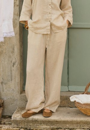 Personne portant un pantalon ample en lin beige et une chemise assortie, avec des ballerines marron, debout sur une marche en pierre près d'une porte verte et d'un panier en osier avec un tissu blanc.
