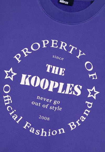 Fioletowy strój z białym okrągłym napisem: "Własność The Kooples od 2008 roku, nigdy nie wychodzi z mody, Oficjalna marka modowa" oraz dwoma ikonami gwiazdek.