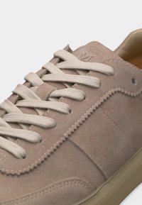 Ανοιχτό μπεζ σουέντ αθλητικό παπούτσι με κρέμ κορδόνια, ζιγκ-ζαγκ ραφή και μπεζ λαστιχένια σόλα, που δείχνει τα πάνω και γλωσσίδα.