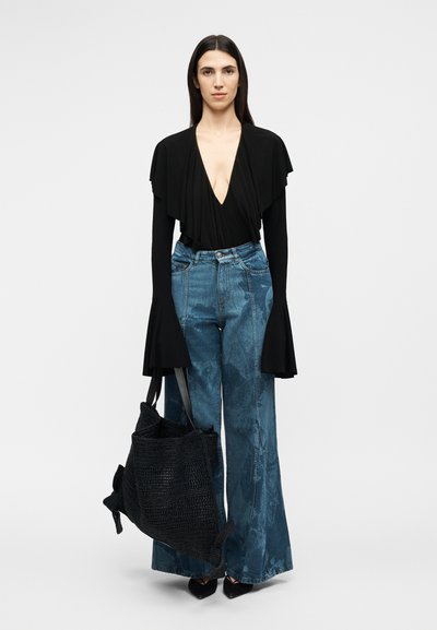 Γυναίκα με μακριά σκούρα μαλλιά, φοράει μαύρο τοπ με βαθύ V και φαρδιά μανίκια, μπλε τζιν με φαρδιά μπατζάκια, μαύρα μυτερά παπούτσια, κρατώντας μεγάλη μαύρη τσάντα tote με υφή.