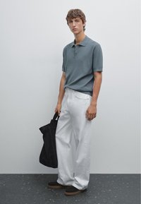 Koszulka polo z krótkim rękawem, prążkowana, w stonowanym niebieskim kolorze, zestawiona z białymi szerokimi spodniami i ciemnymi zamszowymi butami. Modelka trzyma czarną torbę typu tote.