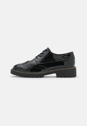 Zwarte lakleren brogueschoen met dikke profielzool, decoratieve perforaties en vetersluiting, zijaanzicht op een witte achtergrond.
