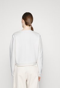 Kvinna med brun hästsvans som bär en enkel vit långärmad tröja och lösa off-white byxor, vänd bort mot en ljus bakgrund.