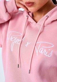 Sudadera de algodón rosa con texto bordado en blanco que dice "Project x Paris". Cuenta con capucha ajustable y puños acanalados, mostrando una textura suave.