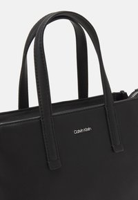 Melna rokassoma ar gludu tekstūru, divām rokturiem un sudraba Calvin Klein logotipu. Aprīkota ar rāvējslēdzēju un minimālisma dizainu.