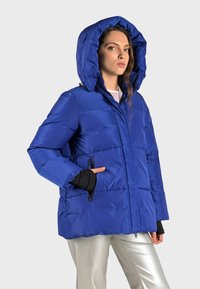 Blauwe pufferjack met capuchon, een ritssluiting aan de voorkant, zijzakken en een gestikt ontwerp. Het materiaal lijkt glanzend en synthetisch.