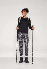 Sieviete pārgājienu aprīkojumā ar rakstainām biksēm un melnu topu, turot pārgājiena nūjas un valkājot mugursomu, smaidot un skatoties labajā pusē.