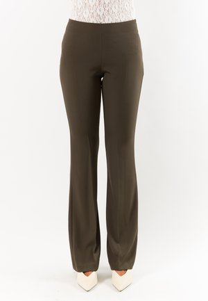 Donna che indossa pantaloni a vita alta a gamba dritta verde oliva scuro e scarpe bianche con punta appuntita su sfondo semplice.