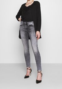 Cardigan negro sobre una blusa negra con encaje, combinado con jeans ajustados grises y zapatos de tacón negros con tiras en el tobillo.