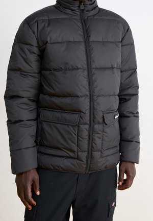 Veste matelassée noire avec fermeture éclair à l'avant et deux poches à rabat, portée par une personne à la peau foncée et portant un pantalon noir.