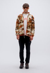 Φλις μπουφάν με πολύχρωμο αφηρημένο σχέδιο, συνδυασμένο με λευκό πουκάμισο με κόκκινο γραφικό βουνό, μαύρο παντελόνι και καφέ παπούτσια πεζοπορίας.