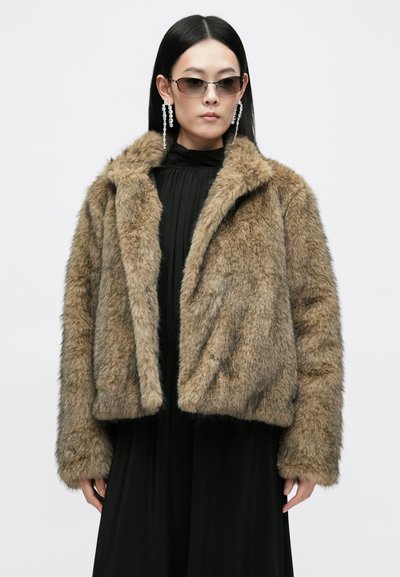 Καφέ ψεύτικη γούνινη ζακέτα με πλούσια υφή, με αυλακωτό γιακά και ανοιχτό μπροστινό μέρος. Φορεμένη πάνω από ένα μαύρο ρούχο, με μακριά μανίκια και τετράγωνη σιλουέτα.