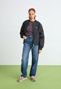 Zwarte gewatteerde bomberjack, gestreepte shirt, hooggemakte blauwe jeans en bruine sneakers met crème accenten. Model staat op groene vloer.