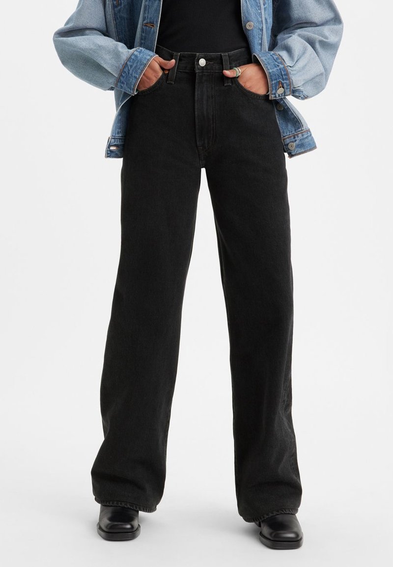 Personne portant un jean noir taille haute à jambes larges, des bottes noires, un haut noir et une veste en jean bleue, les mains dans les poches.
