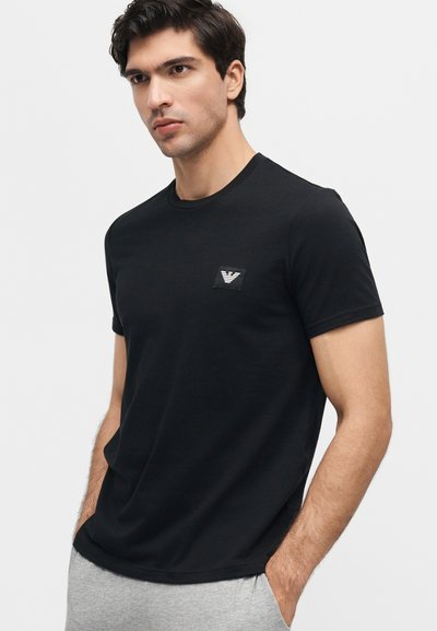 Άνδρας με σκουρόχρωμα μαλλιά φοράει μαύρο κοντομάνικο μπλουζάκι με μικρό λευκό λογότυπο στο στήθος, ανοιχτό γκρι παντελόνι, στέκεται με τα χέρια στις τσέπες.