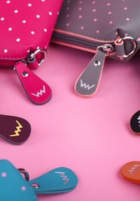 Barevné kožené peněženky s puntíky, které mají odpovídající zipy se šitými cik-cak vzory na růžovém pozadí.