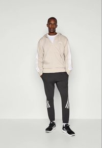 Hanorac crem cu fermoar, având dungi albe pe mâneci, asociat cu pantaloni joggers gri închis cu dungi albe pe laterale și adidași sport negri.