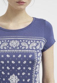 Blå t-shirt med vita paisley- och prickmönster, rund halsringning och korta ärmar, tillverkad av ett mjukt, slätt tyg.