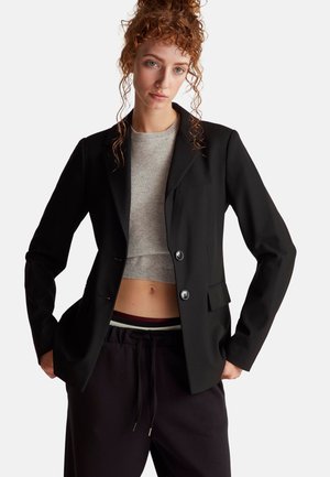 Jonge vrouw met krullend rood haar draagt een zwarte blazer, een grijze cropped trui en zwarte broek met haar handen in de zakken tegen een witte achtergrond.