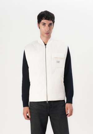 Gilet matelassé blanc avec fermeture éclair à l'avant, doté d'une poche plaquée. Porté sur une chemise bleu marine à manches longues, associé à un jean foncé.