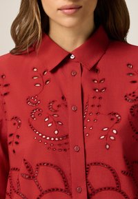 Rotes Hemd mit Knopfleiste und floralem Stickereien, mit Lochmustern und einem strukturierten Kragen. Glatte Textur und mittelschwere Stoff.