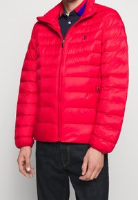 Röd puffjacka med horisontell quiltad design, dragkedjestängning och hög krage. Har en mjuk textur och sidfickor.