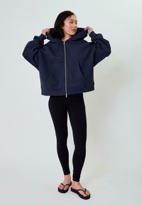 Felpa con zip color navy, dotata di un ampio cappuccio, due tasche frontali e una vestibilità over size. Abbinata a pantaloni legging neri e ciabatte.