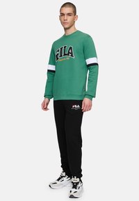 Grön sweatshirt med vita och svarta randiga ärmar, med FILA-logotyp; matchad med svarta sweatpants och FILA-sneakers.