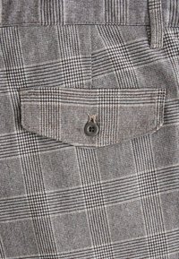 Brown houndstooth patterned fabric with a buttoned pocket. Textured weave with alternating squares and lines, highlighting depth and design intricacy.