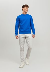 Sweat-shirt bleu en coton, col ras du cou, manches longues, poignets côtelés, associé à un pantalon gris clair et des baskets blanches sur un fond neutre.