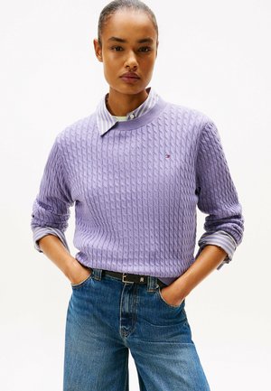 Lilafarbenes Kabelstrickpullover mit rundem Ausschnitt über einem gestreiften Hemd mit Kragen, kombiniert mit hochgeschnittenen blauen Jeans und einem schwarzen Gürtel.