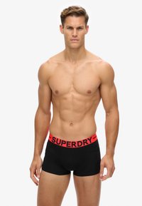 Boxer neri con un ampio elastico rosso che presenta il testo "SUPERDRY." Il tessuto appare liscio con un design aderente.