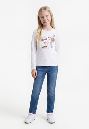 Blond girl standing, wearing a white long-sleeve shirt with printed designs, blue jeans, and white sneakers on a white background.