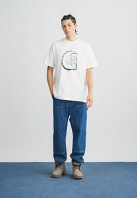 Biele tričko s krátkym rukávom s grafickým dizajnom, kombinované s modrými džínsami a béžovými čižmami. Jednoduchý, voľný strih s okrúhlym výstrihom.