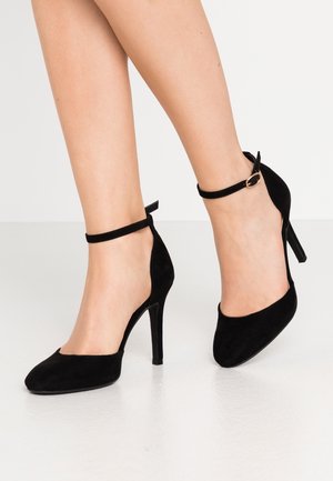 Jambes portant des escarpins en daim noir avec des brides à la cheville et une boucle dorée, posées sur une surface blanche lisse avec un fond uni.
