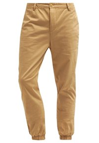 Jasno-brązowe zwężane spodnie z zapięciem na guziki, paskiem i elastycznymi mankietami przy kostkach, wyeksponowane na białym tle.