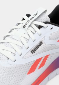 Baltas sportinis batelis su tinkline viršutine dalimi, su paminkštinta apykakle, plokščiomis baltomis raištelėmis ir violetiniu gradientiniu padu. Aiškiai matomas Reebok logotipas.