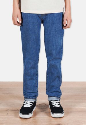 Henkilö, joka on pukeutunut nilkoista käännettyihin sinisiin farkkuihin ja mustiin kengät, joissa on valkoiset nauhat, seisoo puulattialla.