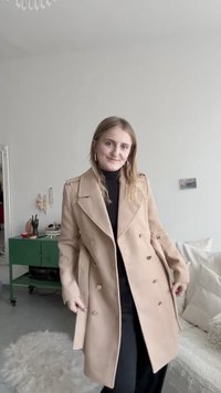 Beige dvouřadový kabát s klopovým límcem, velkými knoflíky a ramenními epolety, zkombinovaný s černým topem a kalhotami.