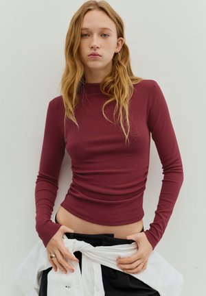 Bordeauxrode top met lange mouwen, een aansluitend silhouet, een zachte textuur en een hoge halslijn, gestyled met een wit overhemd geknoopt in de taille.