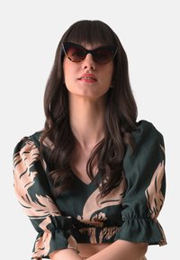 Kattöga solglasögon med svarta rm och sköldpaddsaccenter, bärs av en person i en grön topp med beige blommönster.