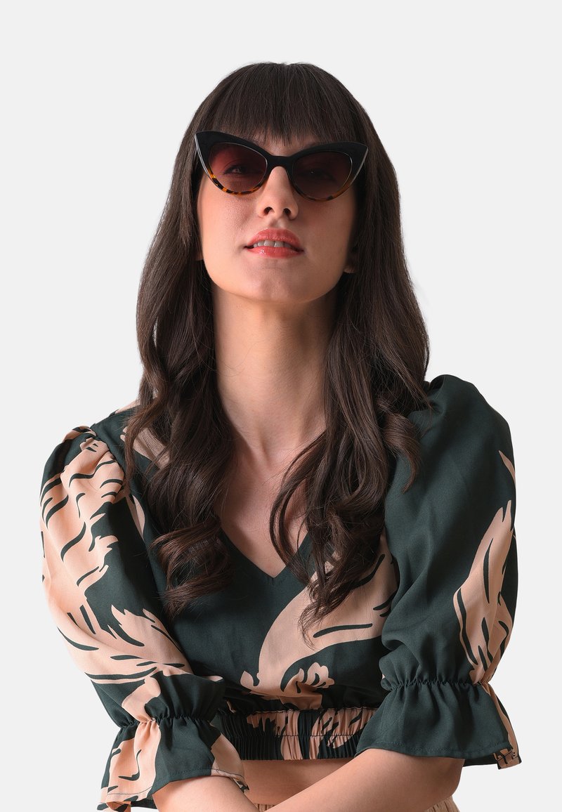 Kattöga solglasögon med svarta rm och sköldpaddsaccenter, bärs av en person i en grön topp med beige blommönster.