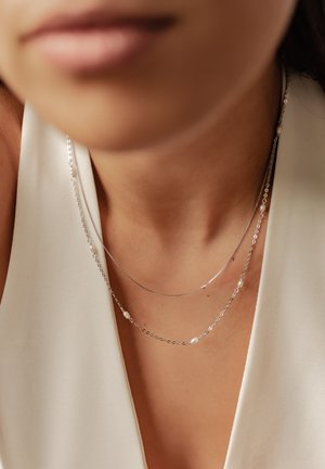 Zwei zarte Silberhalsketten, die übereinander getragen werden, mit einer Mischung aus glatten und strukturierten Gliedern, verziert mit kleinen weißen Perlen.
