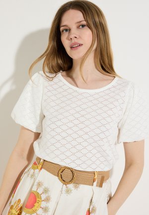 Frau mit langen hellbraunen Haaren, die ein weißes, kurzärmeliges Oberteil mit Lochmuster und einen geblümten Rock mit breitem hellbraunem Gürtel mit runder Schnalle trägt.