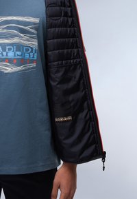 Személy kék grafikás pólóban, részben nyitott fekete dzsekivel, narancssárga cipzárral, amelyen márkázott belső zseb látható.