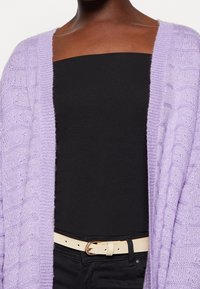 Helllila gestrickter Cardigan mit einem strukturierten Muster, über einem schwarzen Oberteil getragen und mit einem cremefarbenen Gürtel kombiniert.