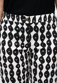 Μέση και άνω μέρος των ποδιών ενός ατόμου που φοράει λευκό παντελόνι με κάθετες μαύρες κυματιστές ρίγες και μικρά μπεζ τετράγωνα μοτίβα, συνδυασμένο με μαύρο τοπ.