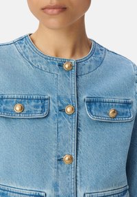 Jasnoniebieska kurtka dżinsowa z okrągłym dekoltem, złotymi detalami, dwiema kieszeniami na piersi i widocznymi stębnowaniami. Materiał to teksturowany dżins.