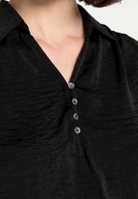 Camicia in tessuto nero, sgualcita, con scollatura a V e cinque bottoni riflettenti leggeri lungo il davanti. Presenta un colletto classico e una texture morbida.