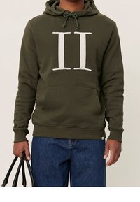 Olivgrön hoodie med en stor vit "II" emblem, dragsko-hoodie, frontficka och ribbade ärmslut. Bärs med blå denim och en svart väska.